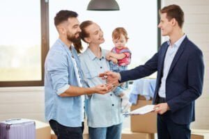 Family of three people take keys new big house from realtor. They smile. Background moving boxes, big window.